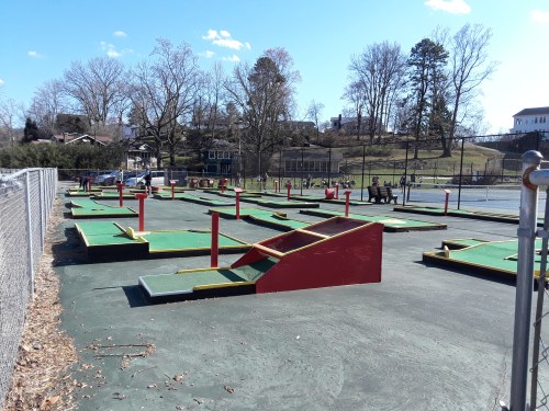 A mini-golf course with colorful obstacles, surrounded by trees and a fence, on a sunny day.