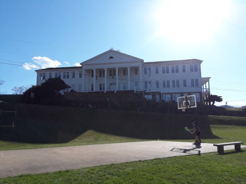 A large white building stands on a hill, with a basketball hoop in the foreground and a child playing nearby.