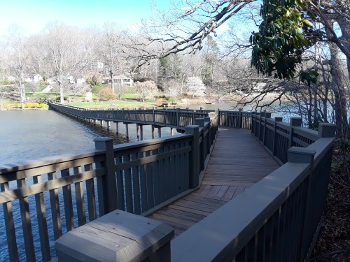 A wooden walkway curves along a lake, surrounded by trees and houses in a serene, natural setting.