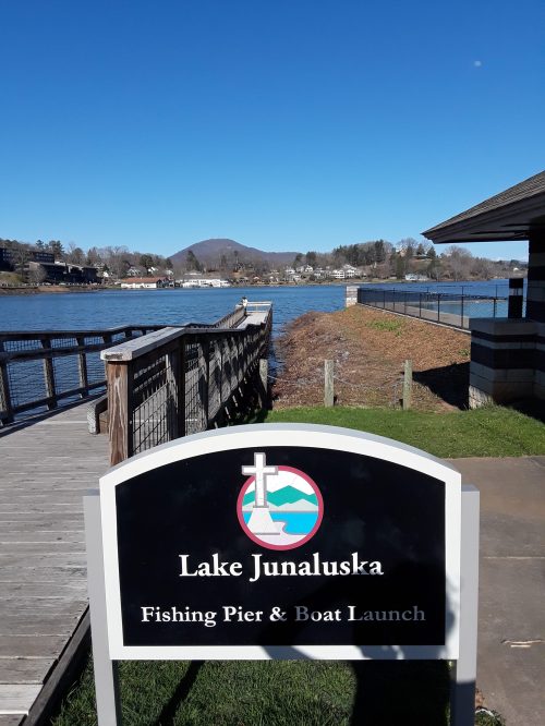 Sign for Lake Junaluska, featuring a fishing pier and boat launch, with a clear blue sky and water in the background.