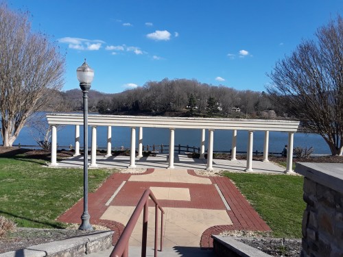 A lakeside pavilion with columns, surrounded by grass and trees, under a clear blue sky. Steps lead down to the area.