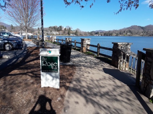 A scenic view of The Rose Walk by a lake, with a sign, trees, and parked cars in the background.