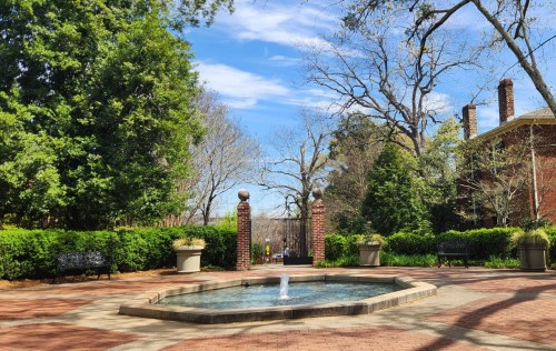 A serene garden scene featuring a fountain, benches, and lush greenery under a clear blue sky.