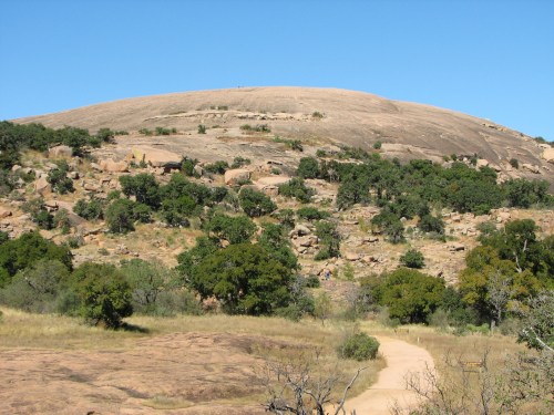 A large, smooth rock formation rises above a grassy landscape, dotted with trees under a clear blue sky.