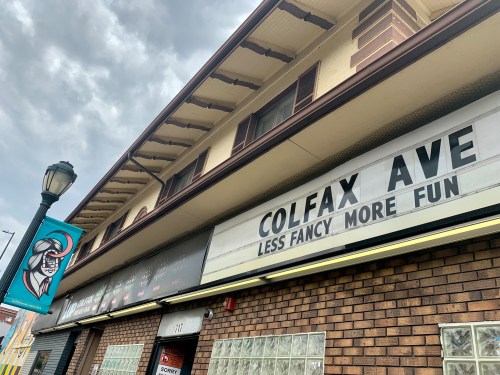 Sign on a brick building reads "Colfax Ave - Less Fancy More Fun," with a decorative banner nearby.