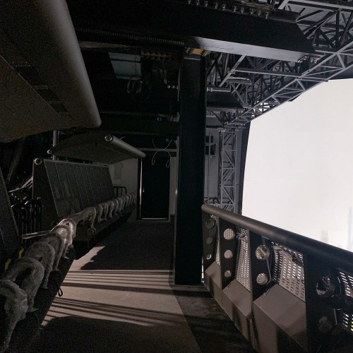 Interior view of a dark, industrial space with seating and a large blank screen.
