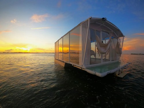 A floating structure with transparent walls on water, set against a vibrant sunset sky.