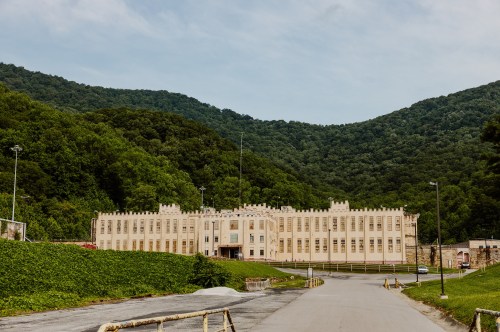 A large, historic building surrounded by green mountains and trees, with a clear sky above.