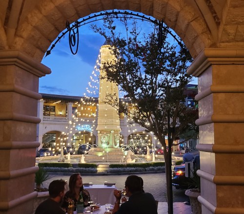 View of a lit fountain and monument through an archway, with diners enjoying a meal in the foreground at dusk.