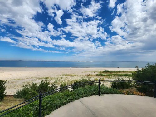 A scenic view of a sandy beach under a blue sky with fluffy clouds, framed by greenery in the foreground.