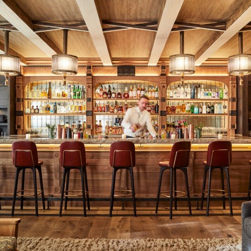 A bartender mixes drinks at a stylish wooden bar with shelves of various bottles and modern lighting.