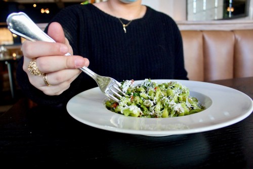 A person holding a fork over a plate of green pasta with vegetables, sitting at a restaurant table.