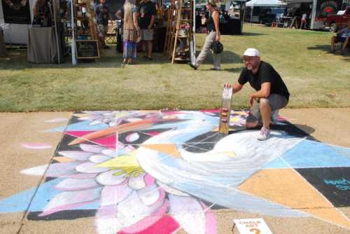A man kneels beside colorful chalk art of a bird on pavement at an outdoor event, with vendors in the background.