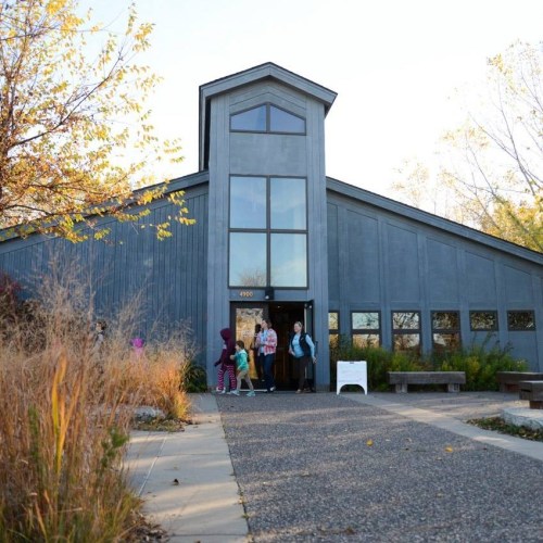 A modern building with large windows, surrounded by greenery, with people entering and exiting.