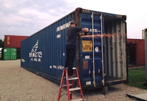 A person on a ladder is cutting a shipping container with a grinder, sparks flying in the air.
