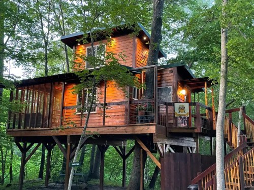 A wooden treehouse nestled among trees, featuring a balcony and warm lights glowing from the windows.