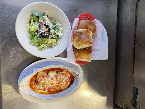 A plate of lasagna with melted cheese, a side salad, and three bread rolls on a metal table.