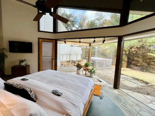 Cozy bedroom with large windows, a bed, and a view of a patio and garden outside. Natural light fills the space.