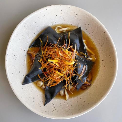 A bowl of black pasta filled with ingredients, topped with crispy orange strips and served in a savory broth.