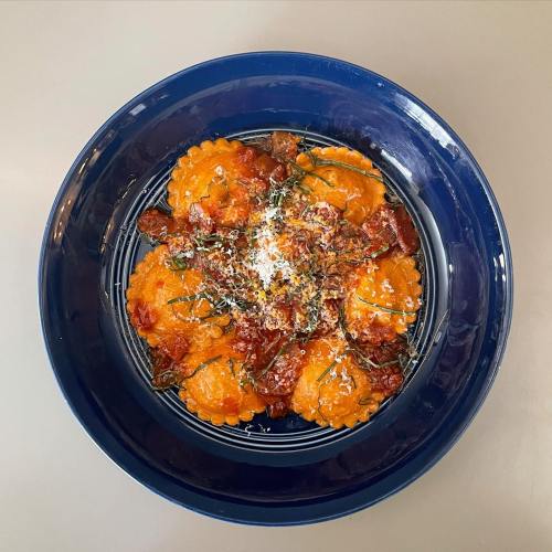 A blue bowl filled with orange ravioli topped with tomato sauce, herbs, and grated cheese on a gray surface.