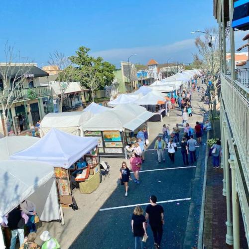 A bustling street market with white tents, vendors, and visitors enjoying the sunny day.