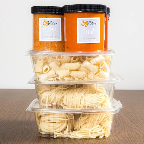 Two jars of pasta sauce on top of three containers of different types of pasta, arranged on a wooden surface.