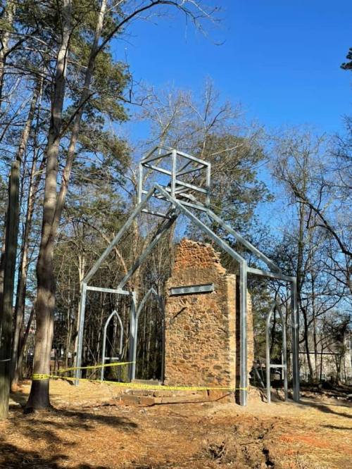 A partially constructed structure with a stone wall, surrounded by trees and caution tape, under a clear blue sky.
