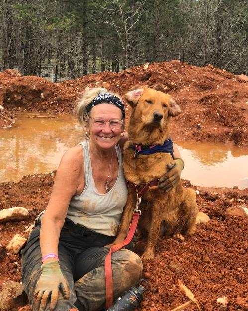 A smiling woman in a tank top sits in muddy ground with a golden retriever beside her, both looking happy.