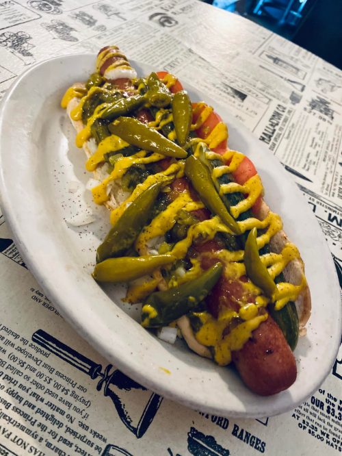 A hot dog topped with mustard, green peppers, and tomatoes, served on a white plate on a newspaper-print table.