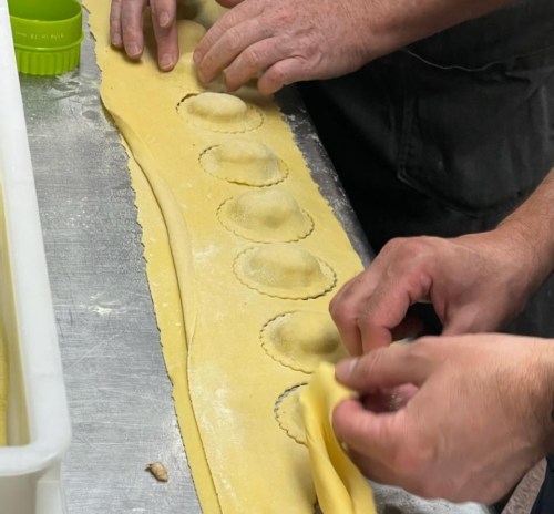 Two hands are shaping ravioli on a sheet of pasta, with flour scattered on the surface.