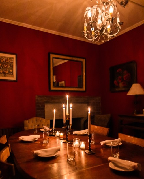 A cozy dining room with a wooden table set for dinner, candles lit, and warm red walls illuminated by a chandelier.