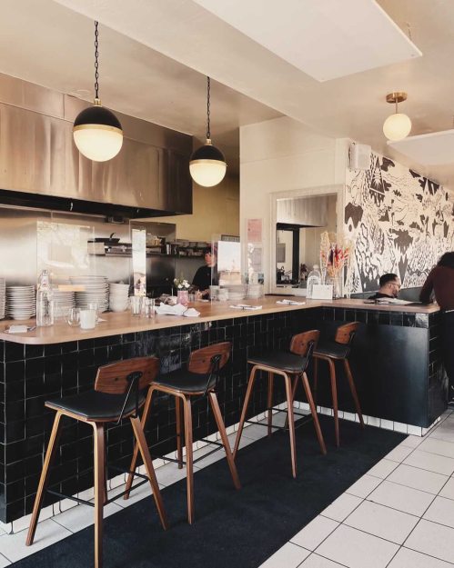 A modern café interior featuring a sleek bar, wooden stools, and stylish lighting, with a floral mural in the background.