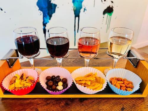 A wooden tray holds four glasses of wine and four bowls of snacks, including nuts, chips, and chocolate.