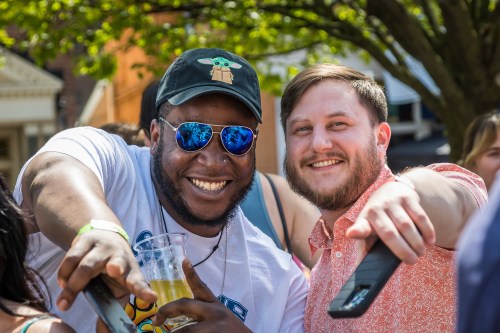 Two smiling men point at the camera, one wearing sunglasses and a cap, holding a drink, in a sunny outdoor setting.