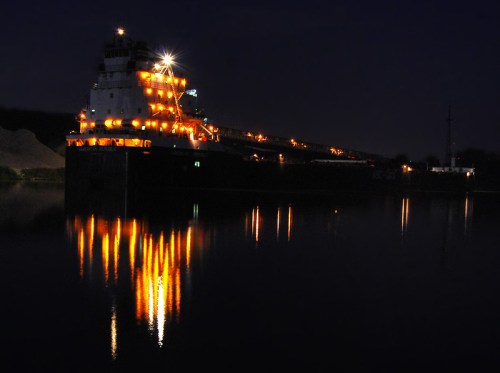 A large ship illuminated at night, reflecting lights on calm water, with a dark background and minimal shoreline visible.