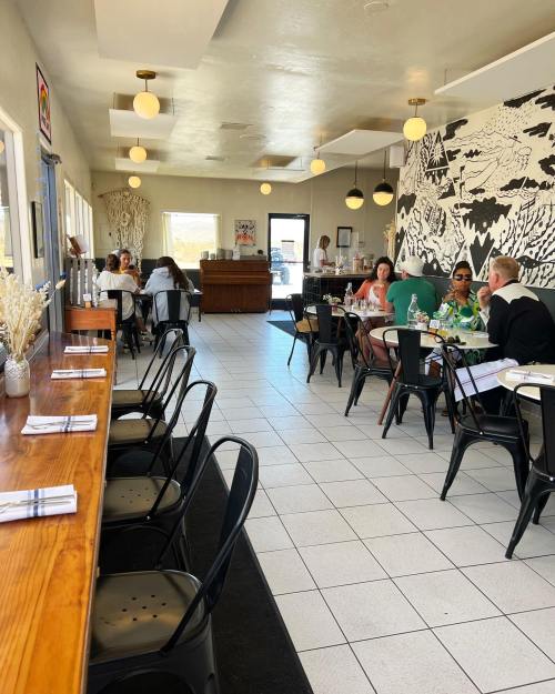 A bright restaurant interior with tables, chairs, and a mural, featuring diners enjoying their meals.