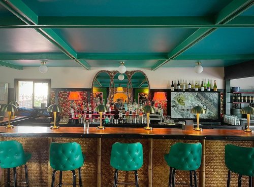 A stylish bar with green stools, gold lamps, and a vibrant decor featuring mirrors and colorful artwork.