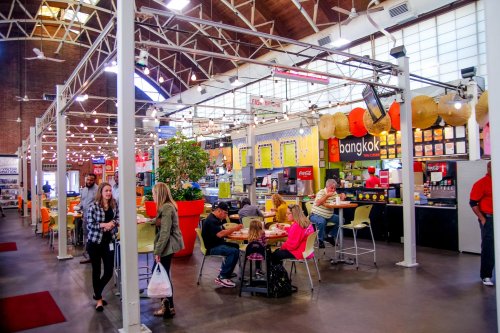 A vibrant food hall with colorful stalls, people dining, and a lively atmosphere under bright lights.