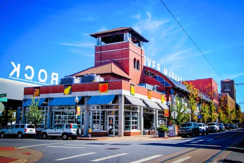 A vibrant market building with a tower, surrounded by trees and parked cars, under a clear blue sky.