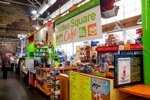 Colorful café interior with a menu sign, food displays, and a self-service area in a vibrant market setting.