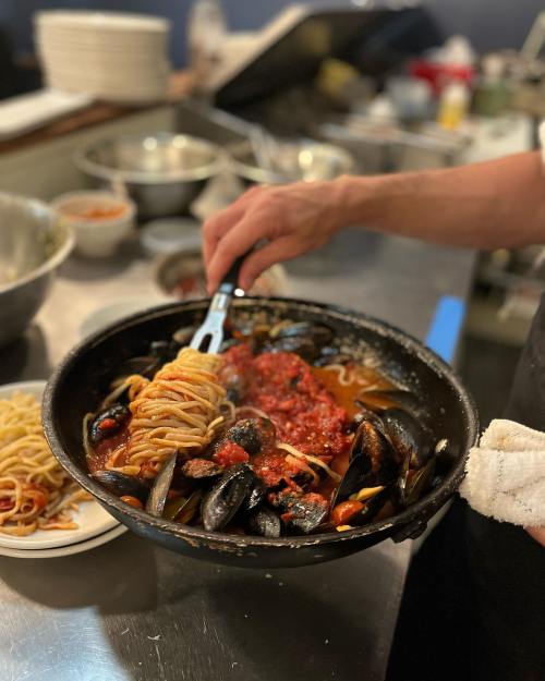 A chef's hand twirls spaghetti with mussels and marinara sauce in a large skillet, surrounded by kitchen utensils.