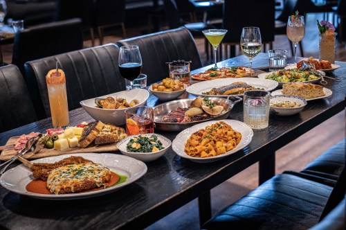 A table filled with various dishes, including pasta, seafood, salads, and drinks, in a stylish restaurant setting.