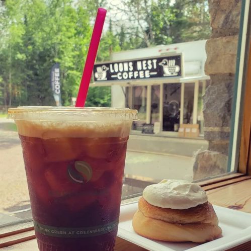 A cold drink with a pink straw next to a pastry, viewed through a window with a coffee shop in the background.