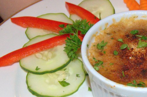 A ramekin of creamy dish garnished with herbs, surrounded by cucumber slices and red bell pepper strips.