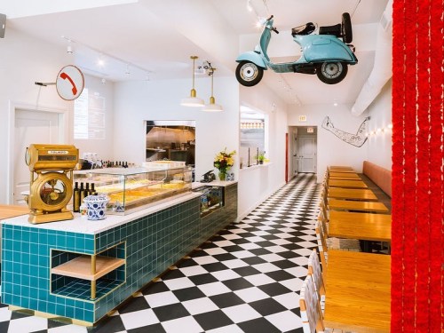 A modern café interior with a blue scooter on the ceiling, checkered floor, and wooden tables along the walls.