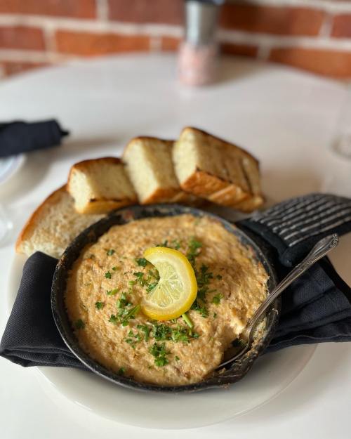 A bowl of creamy dip garnished with lemon and herbs, served with slices of toasted bread on a white table.