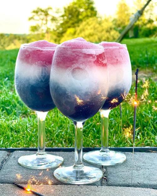 Three colorful frozen drinks in elegant glasses, with a sparkling sparkler in front, set against a green outdoor background.