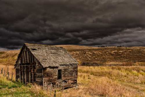 A weathered wooden cabin stands in a grassy field under a dramatic, dark stormy sky.