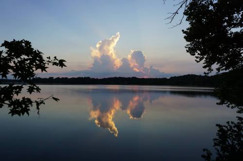 A serene lake at sunset, reflecting colorful clouds and surrounded by trees.