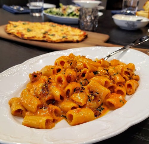 A plate of pasta in a creamy orange sauce, garnished with herbs, with pizza and salad in the background.